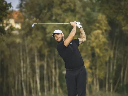 Golfer in Schwarz beim Abschlag auf einem Fairway zwischen hohen Bäumen