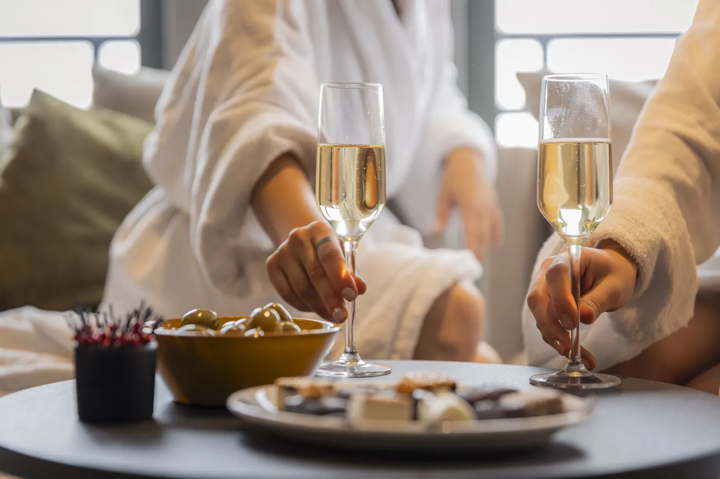 Zwei Hände mit Champagnergläsern über einem Tisch mit Oliven und Pralinen in der Wellness-Lounge